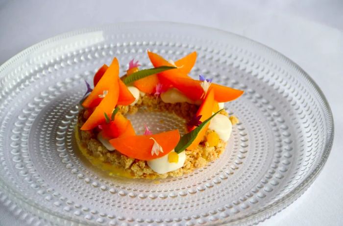 A glass plate showcases a circular dessert made of orange-fleshed fruit, garnished with herbs and edible flowers, resting on dollops of cream and a crumble base.