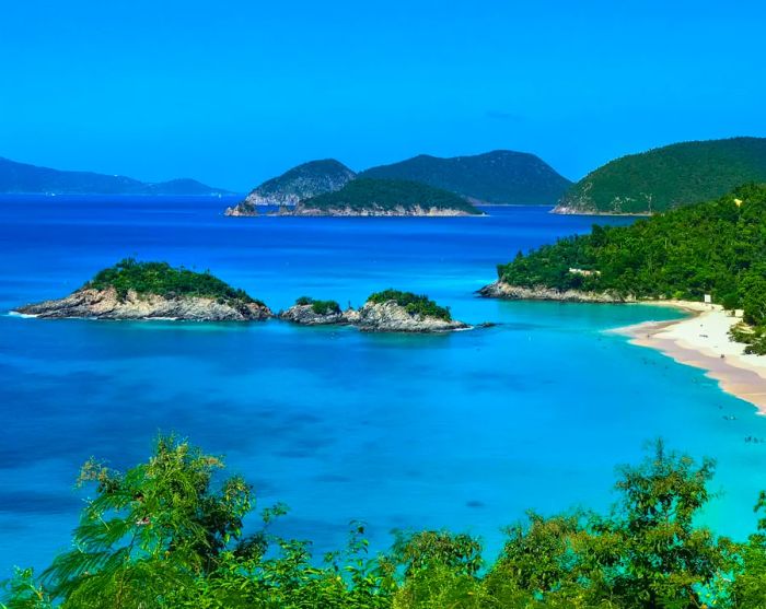 A pristine blue bay with white sands and offshore islets; green mountains rise in the background.