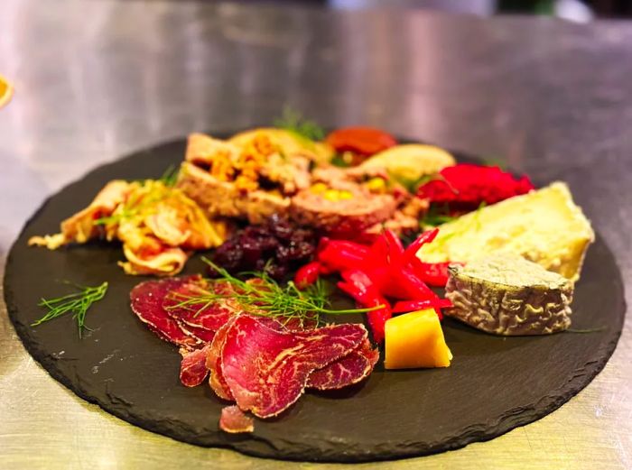 A dark platter showcasing an array of charcuterie and cheese garnished with herbs and peppers.