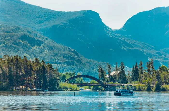 View from the lake: a pontoon boat near an arched bridge, surrounded by evergreen trees and towering green mountains.