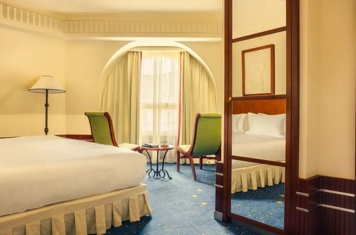 A bright guest room at Hôtel des Grands Voyageurs, featuring blue carpeting and two green chairs beside a large window.