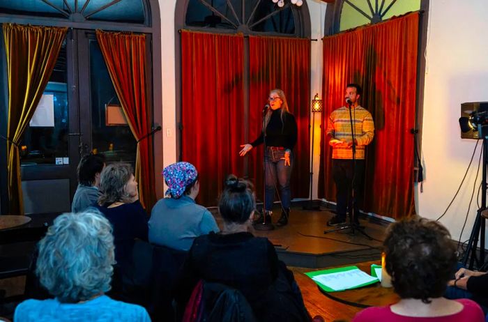 Two artists are engaging the audience with microphones on a stage adorned with vibrant red curtains at Story Parlor.