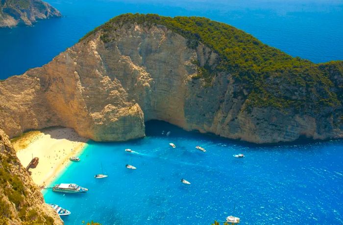 Aerial view of a secluded beach featuring a rusted shipwreck, bordered by cliffs and stunning blue waters, with several small white boats nearby.