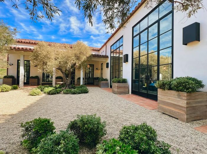 Courtyard at The Ranch Malibu, shaded by trees; a white single-story building featuring a wall of windows