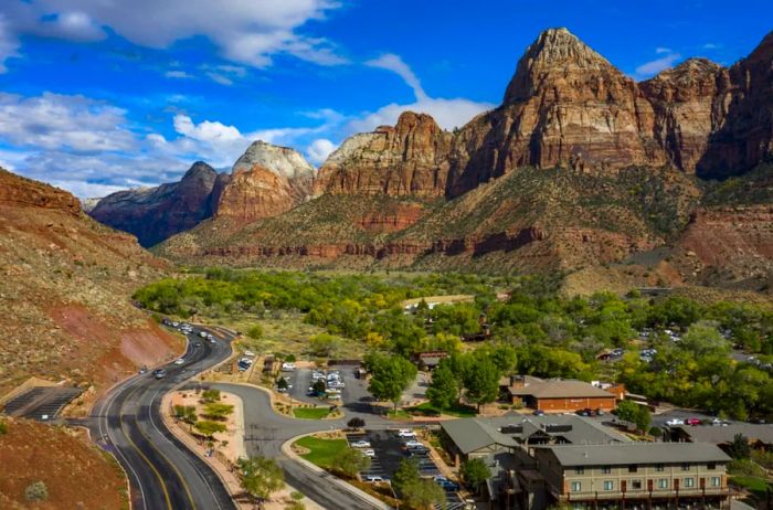 An aerial view showcases low buildings adjacent to a winding road, framed by red-rock formations beneath a bright blue sky adorned with fluffy clouds.