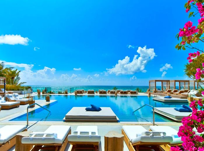 The outdoor swimming pool at 1 Hotel South Beach is surrounded by white benches, with vibrant pink and purple bougainvillea flowers framing the right side of the scene.