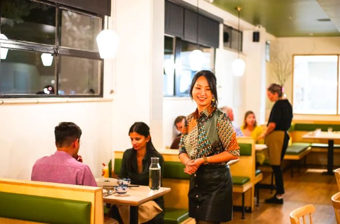 A woman stands in the center of a restaurant’s bright dining area, surrounded by diners at cozy booths.