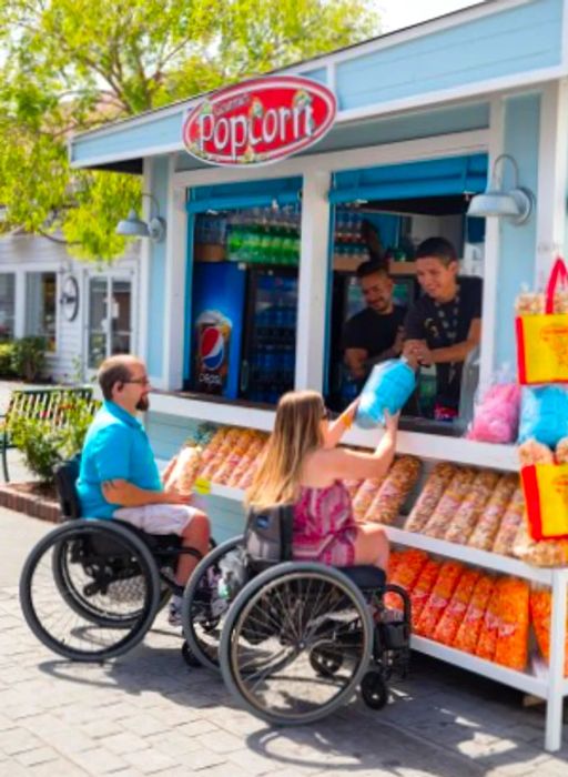 Two individuals in wheelchairs enjoying cotton candy at an outdoor food stand.