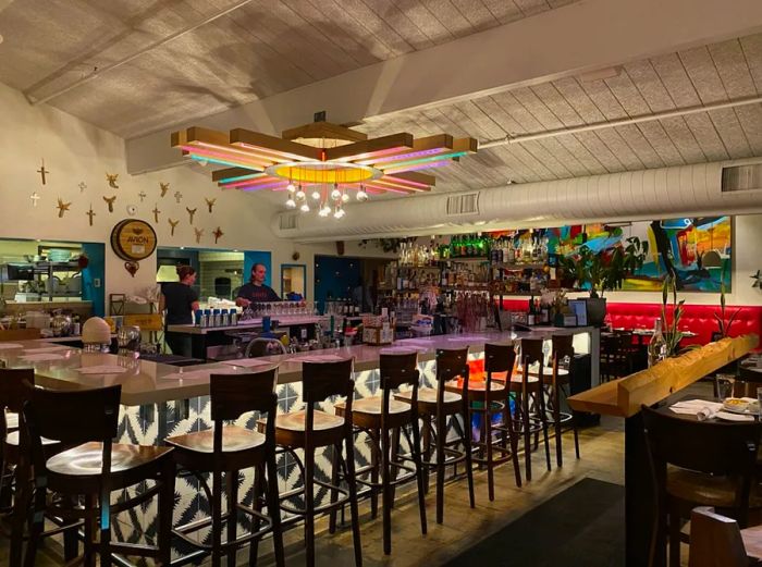 A bar featuring decorative tiles, a row of unoccupied brown high-top seats, a red banquette along the back wall, and a cross-shaped light fixture overhead.