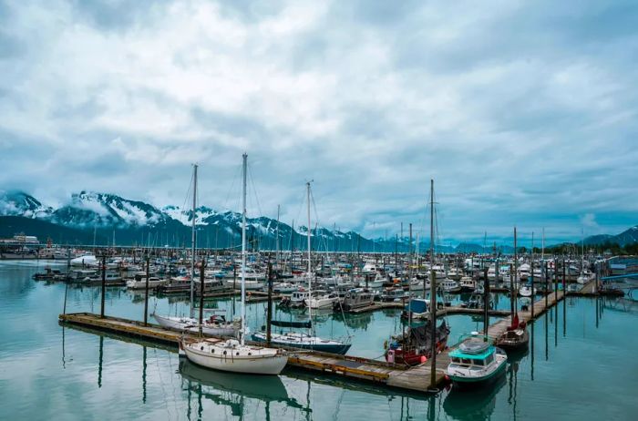 Boats are anchored in a harbor, with mountains and dramatic cloudy skies in the background.