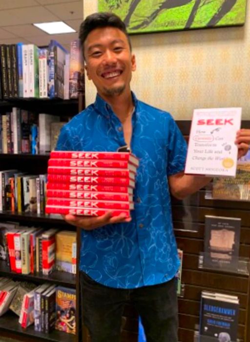 In a bookstore, Scott Shigeoka poses with copies of his book 'Seek,' which features a white cover and a red spine.