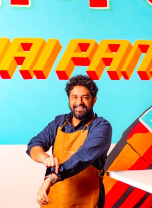 Chef and entrepreneur Meherwan Irani, donning an apron and rolling up his sleeves, stands before a vibrant mural.