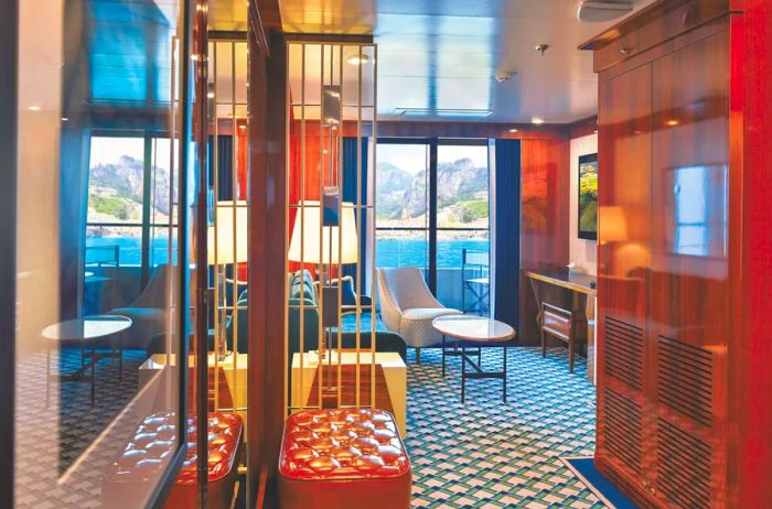 A glimpse of the sitting area in the Navigator Suite aboard the 'World Traveller,' featuring blue-and-white patterned carpeting, a leather bench, a sofa, an armchair, and a balcony.