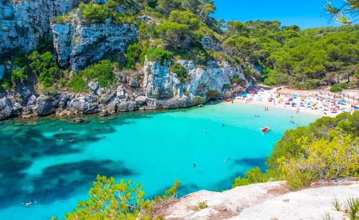 A hidden cove featuring bright blue-green waters and a sandy beach adorned with colorful umbrellas, with a few swimmers enjoying the water.