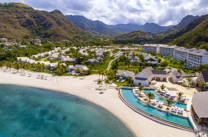 Aerial view showcasing the beach and expansive resort, featuring a curved pool by Buccament Bay with verdant mountains in the backdrop.