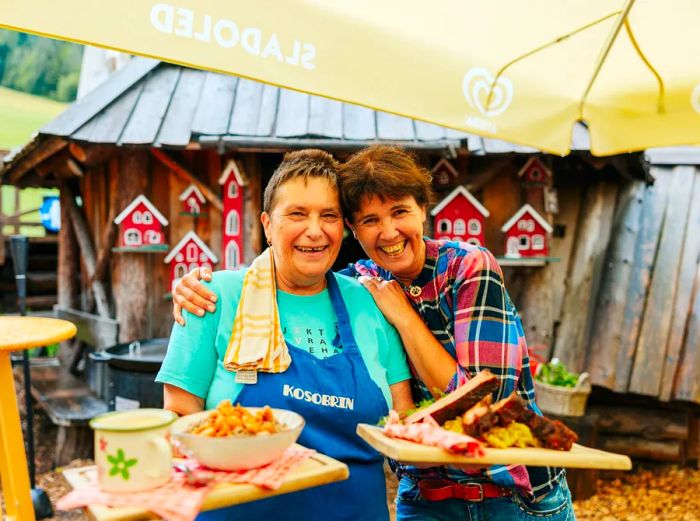 Two women outside, one carrying two wooden trays filled with food