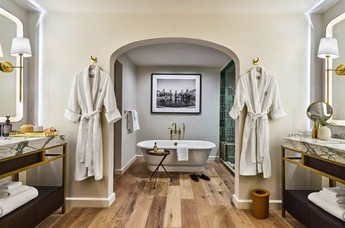 A bathroom featuring a deep soaking tub, accompanied by hanging robes and marble vanities in the foreground.