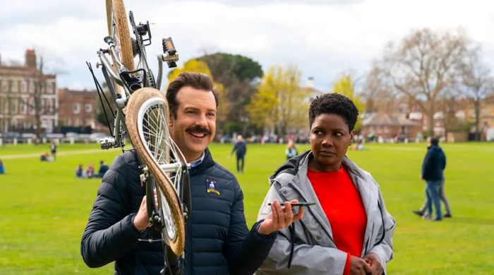 Ted Lasso, with a damaged bike slung over his shoulder, strolling through a park alongside his doctor.