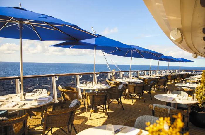 An outdoor dining area on the 'Seven Seas Splendor' featuring several tables and chairs beneath blue umbrellas, offering breathtaking views of the sea.