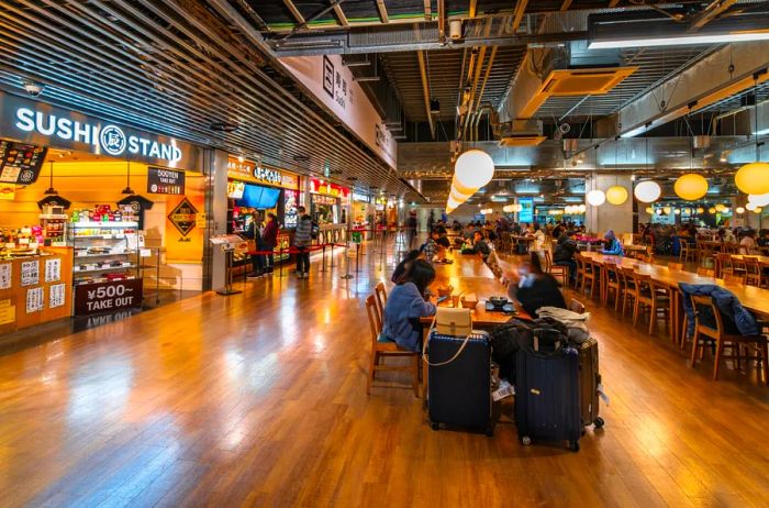 A dining area at Narita International Airport features a sushi stand prominently in the foreground, with travelers seated at long tables, accompanied by their luggage.