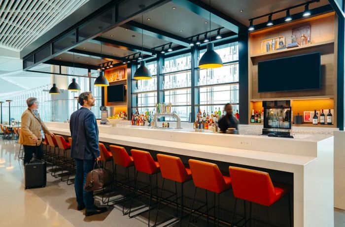 Two men are standing at a long bar, waiting for the bartender to serve them in the United lounge at Newark Airport.