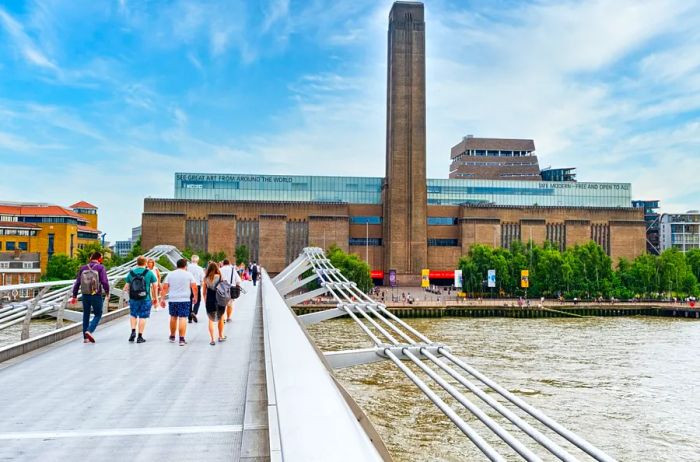 A sleek metal pedestrian bridge spans the River Thames, leading to the Tate Modern, a massive brick structure crowned with a tall rectangular tower.