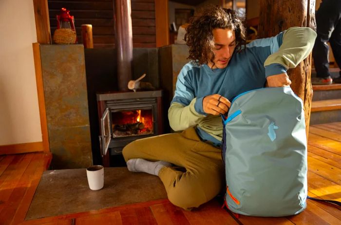 A man relaxes in front of a fireplace indoors, reaching into a light-blue Cotopaxi backpack.