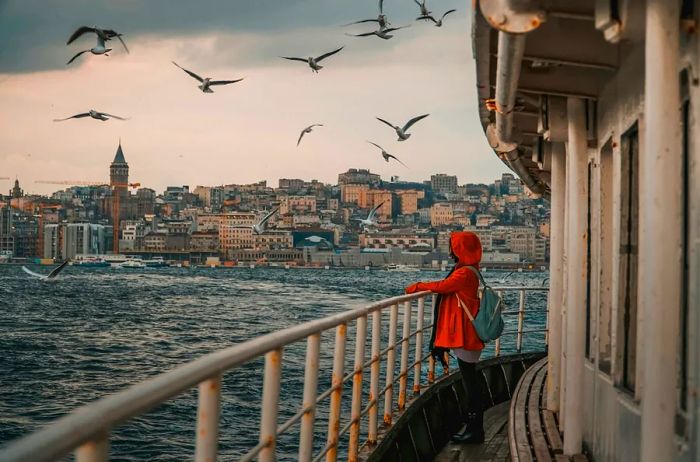 Individual wearing an orange hooded jacket standing on a boat, with the Istanbul skyline in the backdrop and seagulls soaring above.