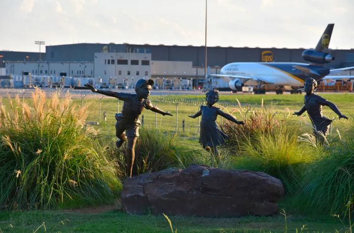 A dark metal sculpture depicting three children running, located outdoors at DFW's Founder's Plaza observation area.