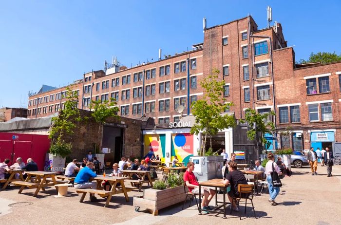 Groups of people enjoying drinks and socializing at picnic tables outside a bar, with a multi-story brick building in the background