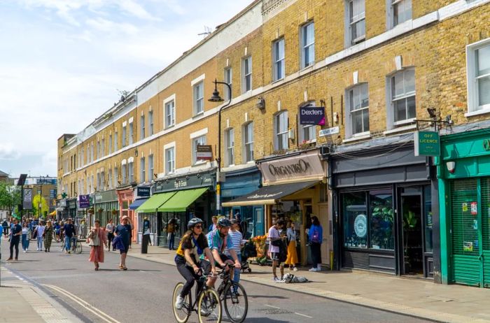 A few cyclists pedaling down the middle of a London street that is lined with shops, with pedestrians visible in the distance.