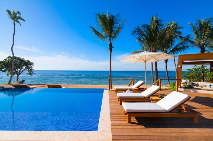 The pool area offers stunning views of the Caribbean, featuring white lounge chairs set on a wooden deck at Casa de Campo Resort and Villas in La Romana, Dominican Republic.