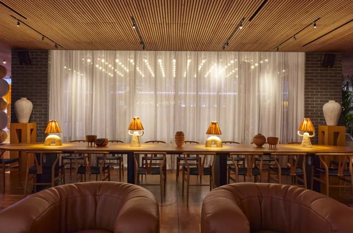 A long table with chairs, topped with small lamps, alongside two tan curved armchairs in the lobby of One Hundred Shoreditch hotel.