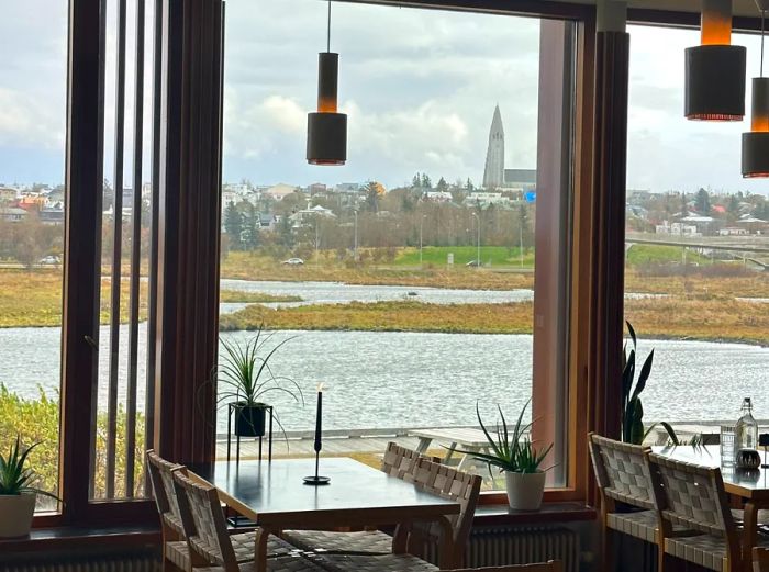 Two vacant restaurant tables beside expansive windows at Nordic House in Reykjavík, overlooking the water and cityscape in the distance.