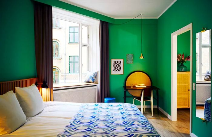 A triple room at the Coco Hotel in Copenhagen features vibrant green walls, a compact desk, and a double bed dressed in a blue-and-white duvet cover.