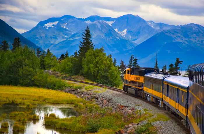 A blue and gold train glides past a tranquil pond with mountains looming in the background.