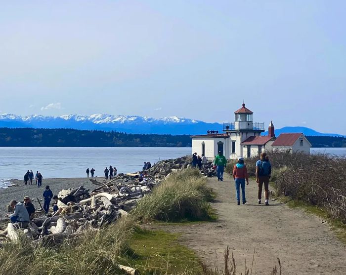 A charming white lighthouse with a red roof stands in Discovery Park, with snow-capped mountains and water in the background, while a few people stroll along the trail in the foreground.