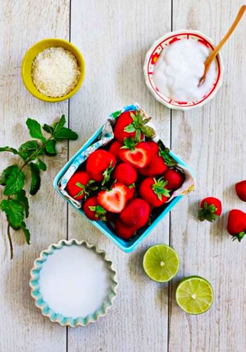 Strawberries, limes, coconut milk, coconut yogurt, mint leaves, and salt displayed on a wooden table.