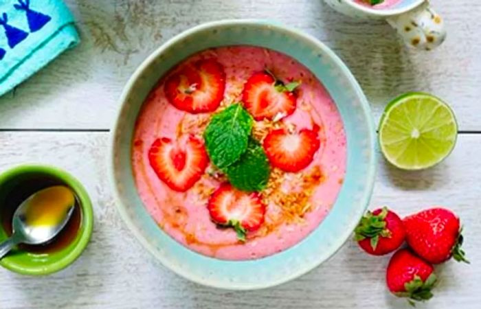 A bowl of strawberry coconut soup adorned with mint leaves, toasted coconut, slices of strawberry, and a drizzle of coconut nectar.