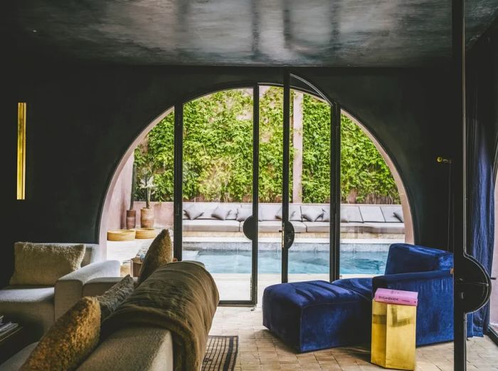 A sleek, modern room featuring a green couch and blue armchair, leading through an arched door to a poolside terrace.