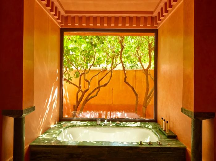 The bathroom at Amanjena in Marrakech features a luxurious soaking tub with a view of a lush garden and trees beyond.