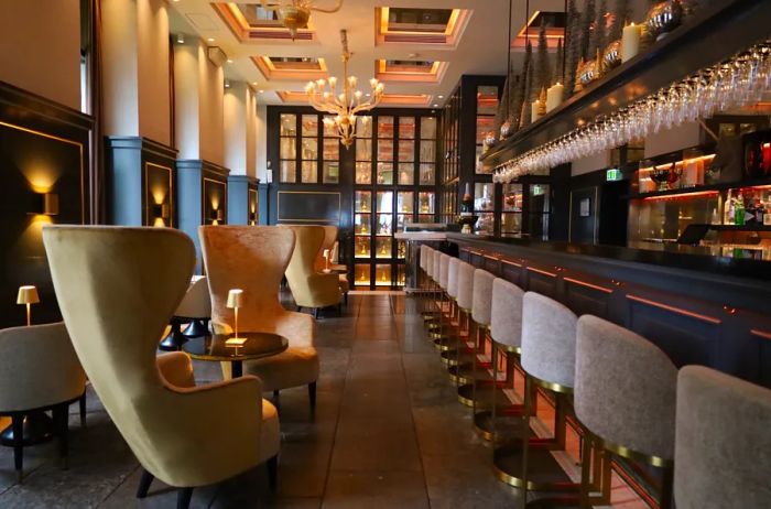 Gray upholstered bar stools line the elongated Balthazar Champagne Bar in Hotel d'Angleterre, complemented by oversized armchairs and cocktail tables positioned nearby.