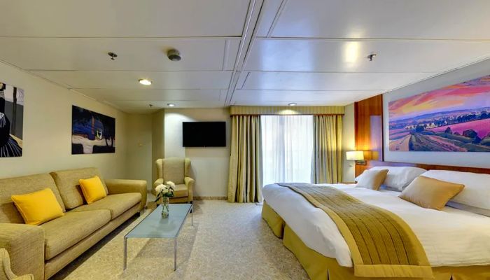 A Superior Suite aboard the upcoming Villa Vie Odyssey residential cruise ship, featuring a king-sized bed, a couch, an armchair, and curtains, along with a private balcony.