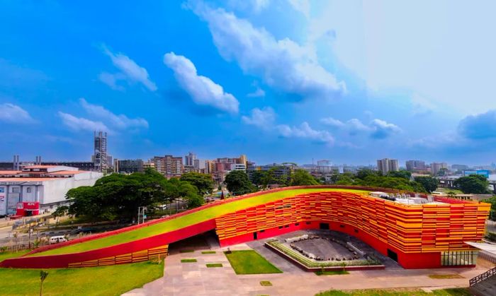 A vibrant, sloped building with a green roof, set against an urban skyline backdrop