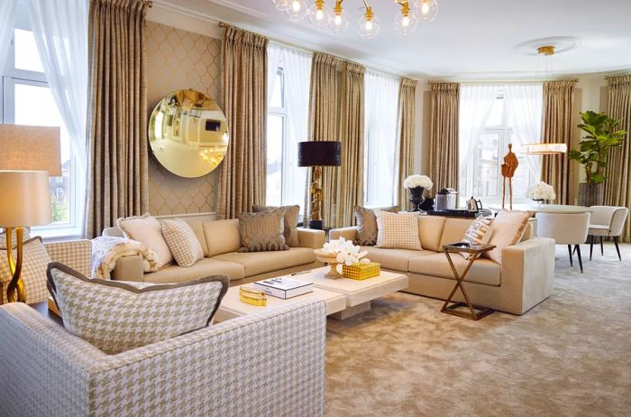 The living area of a d'Angleterre Suite features three beige sofas, floor-to-ceiling windows adorned with drapes, a fiddle leaf fig tree, and a large round mirror.