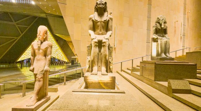 Three ancient Egyptian statues stand along a grand staircase inside a museum, surrounded by sand-colored walls and floors.