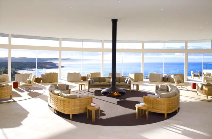 A sweeping white Great Room featuring floor-to-ceiling windows, empty chairs oriented towards the ocean, and a striking black suspended fireplace at its center.