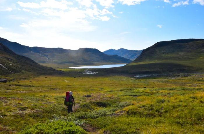 A female hiker traverses a breathtaking landscape featuring mountains, azure lakes, rocks, and tundra.