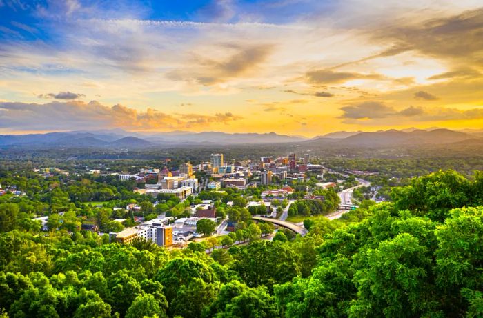 A panoramic view of Asheville's downtown framed by lush trees, mountains, and a blue sky dotted with clouds.