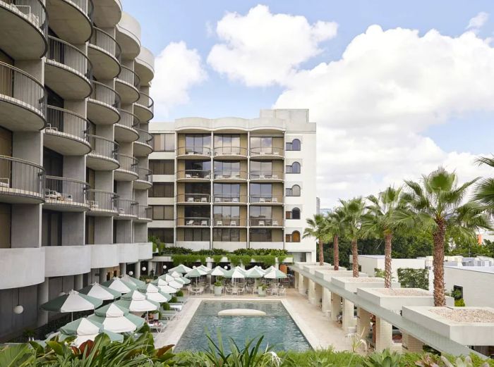 The exterior of the five-story Calile Hotel is white, featuring balconies that overlook the swimming pool and a row of palm trees.